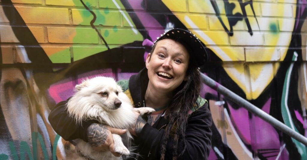 Woman smiling wide and holding her white dog in front of mural.