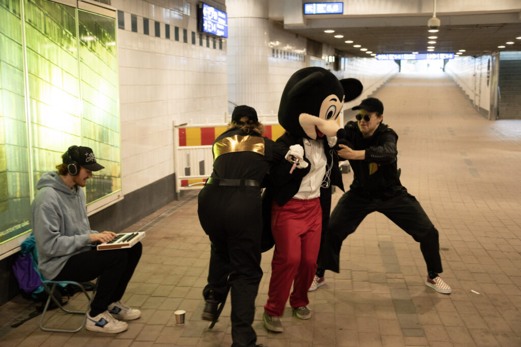 Two security guards try to catch Mickey Mouse, who is dressed in a tailcoat in the subway tunnel. A man sits on the edge of a tunnel and plays a synthesizer with headphones on and smiles.
