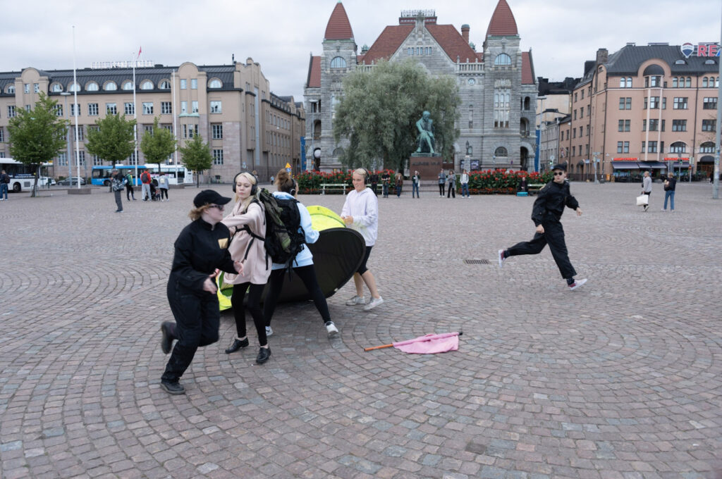 People in the market square. The participants of the show hold the tent in their hands, two guards run around. Participants have headphones. National Theater in the background.